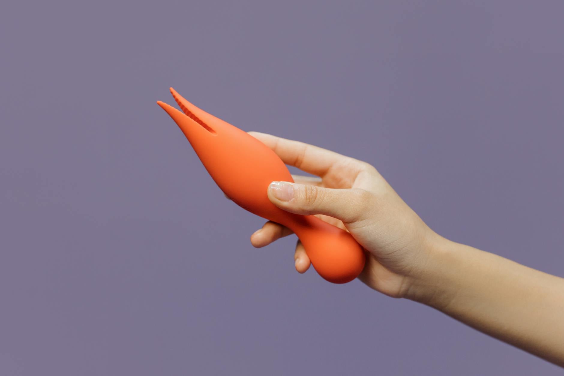Eine Hand hält einen orangefarbenen Vibrator gegen einen minimalistischen violetten Hintergrund